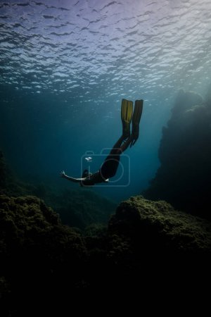 Foto de Cuerpo completo de buzo masculino irreconocible en traje de neopreno y máscara de snorkel flotando en la espalda en aguas profundas oscuras cerca de coral - Imagen libre de derechos