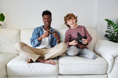 Photo for Full body smiling multiracial male and teenage boy in casual clothes playing video game with joysticks while sitting with legs crossed on soft couch - Royalty Free Image