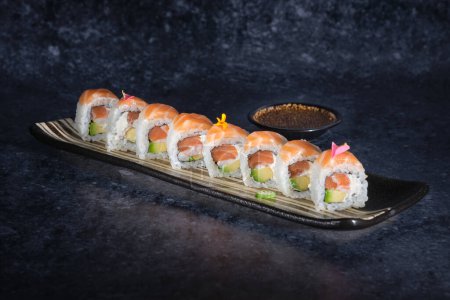 Foto de Fila de frescos rollos de sushi de California con salmón y flores servidas en bandeja de mármol sobre fondo oscuro cerca de un tazón - Imagen libre de derechos