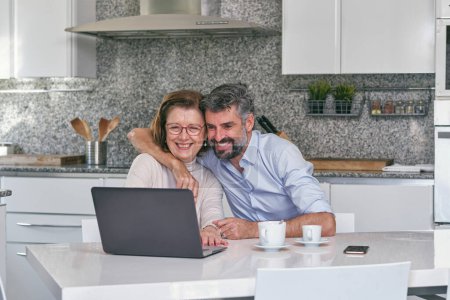 Téléchargez les photos : Homme souriant étreignant femme âgée tout en regardant l'écran d'ordinateur portable et en riant dans la cuisine moderne en marbre à la maison - en image libre de droit
