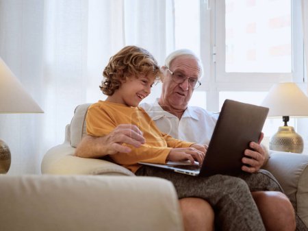 Téléchargez les photos : Délicieux homme âgé avec petit-fils tapant sur ordinateur portable moderne tout en étant assis ensemble dans un fauteuil dans le salon léger à la maison - en image libre de droit