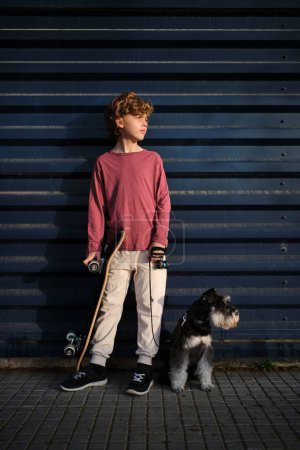 Photo for Full length little skater with skateboard and loyal dog on leash looking away while resting near building wall - Royalty Free Image