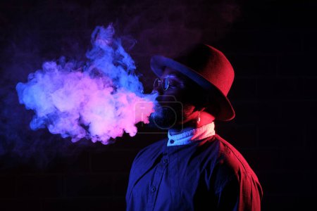Photo for Side view of bearded African American man in stylish hat and sunglasses smoking in dark room with vape - Royalty Free Image