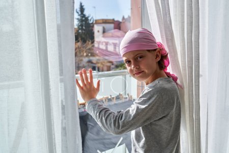 Photo for Side view of kid in pink headband touching glass on balcony door and looking at camera in daylight - Royalty Free Image