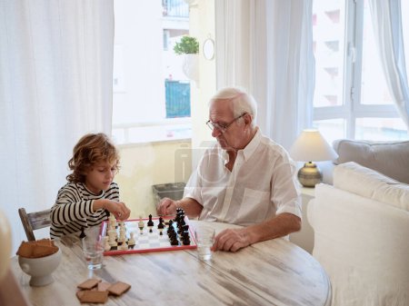 Foto de Abuelo y nieto se unen mientras juegan al ajedrez mientras están sentados en la mesa de madera con galletas en la acogedora sala de estar en casa - Imagen libre de derechos