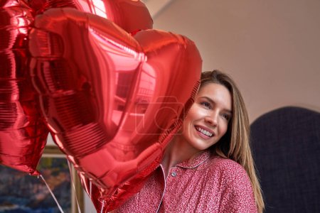 Foto de Mujer feliz con un ramo dotado de globos de papel de aluminio en forma de corazón rojo sonriendo y mirando hacia otro lado mientras celebra el Día de San Valentín - Imagen libre de derechos
