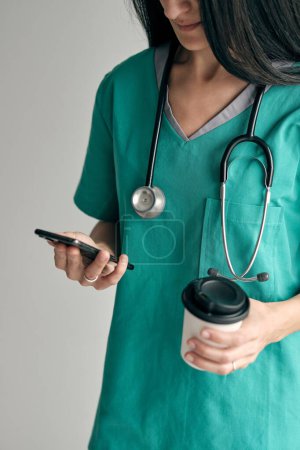 Téléchargez les photos : Cultivez chirurgien femme méconnaissable en uniforme médical vert avec boisson chaude pour aller messagerie texte sur téléphone mobile sur fond clair - en image libre de droit
