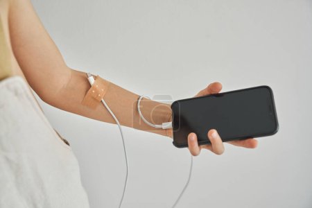 Photo for Crop digital addicted kid with white wire connected to arm with adhesive plaster demonstrating smartphone while standing against gray background - Royalty Free Image
