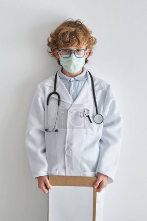 Téléchargez les photos : Enfant en lunettes et peignoir médical avec masque jetable et feuille de papier vide regardant la caméra sur fond blanc - en image libre de droit