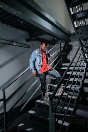 Photo for Young black male in street style clothes thoughtfully standing on staircase in building and looking down - Royalty Free Image