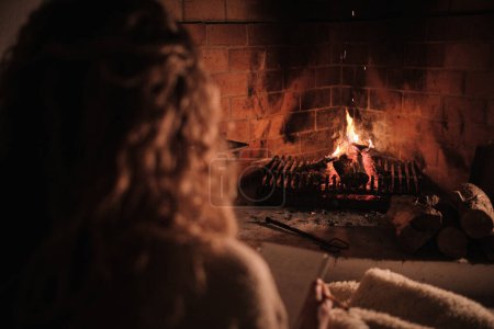 Téléchargez les photos : Vue arrière d'une femme méconnaissable reposant sur le sol près d'une cheminée en feu et lisant une histoire intéressante dans un salon confortable à la maison - en image libre de droit