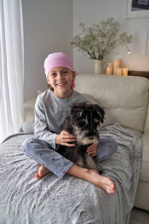 Photo for Full body of cheerful cheerful kid in pink bandana symbol of awareness and fight against breast cancer sitting with crossed legs and stroking fluffy Miniature Schnauzer dog on couch at home - Royalty Free Image