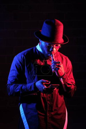 Photo for Black male model in trendy outfit with hat smoking e cigarette and chatting via phone on dark background in neon lighting - Royalty Free Image
