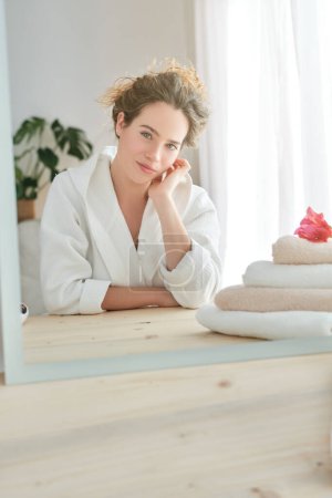 Téléchargez les photos : Charmante femme en peignoir regardant la caméra assise à table près du miroir et des serviettes pendant la routine matinale à la maison - en image libre de droit