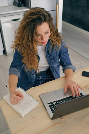 Téléchargez les photos : Entrepreneur féminin concentré assis à table et tapant sur netbook tout en faisant des recherches pendant le travail sur le projet à distance et en prenant des notes dans un bloc-notes dans la cuisine à la maison - en image libre de droit