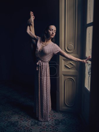 Foto de Bailarina de cuerpo entero en vestido largo de pie contra la puerta en habitación oscura mientras sostiene la pierna y cierra los ojos - Imagen libre de derechos