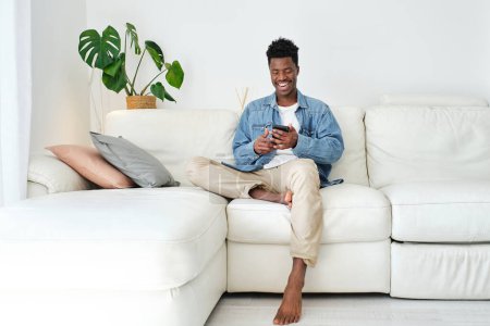 Photo for Happy African American male text messaging on cellphone while resting on couch in living room - Royalty Free Image