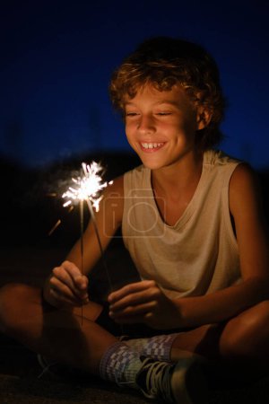 Joyful preteen boy sitting with glowing sparklers at night and celebrating festive occasion