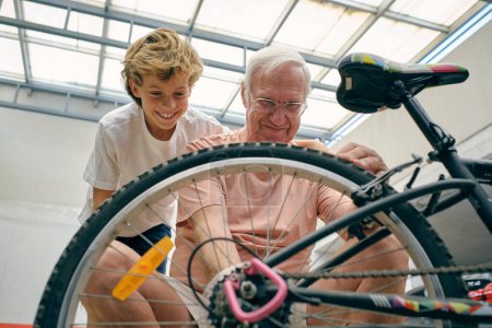 Téléchargez les photos : Grand-père positif avec les cheveux gris dans les lunettes de réparation vélo avec le soutien de garçon debout près - en image libre de droit