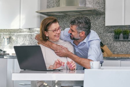 Téléchargez les photos : Femme âgée souriante et homme mûr se regardant et s'embrassant tout en étant assis à table avec netbook dans la cuisine moderne en marbre à la maison - en image libre de droit