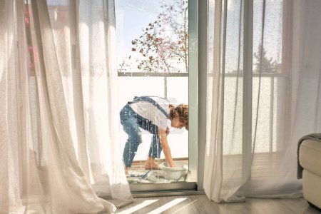 Photo for Side view of boy with curly hair standing on towel and rinsing rag in basin while doing household chores and washing windows on balcony at home - Royalty Free Image