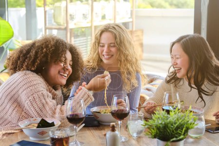 Téléchargez les photos : Groupe de femmes diverses souriant et traînant au restaurant tout en mangeant de délicieux ramen et en buvant du vin - en image libre de droit