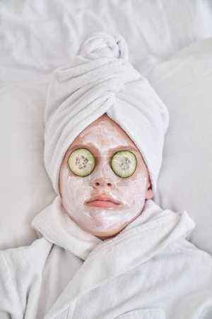 Téléchargez les photos : Vue du dessus du garçon avec masque facial et tranches de concombre portant turban serviette et peignoir relaxant sur lit doux pendant les procédures de beauté dans la chambre à coucher à la maison - en image libre de droit