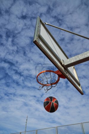 Foto de Bajo ángulo de la bola que baja del aro de baloncesto con la red colocada en el tablero contra el cielo azul nublado - Imagen libre de derechos