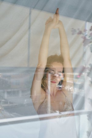 Photo for Through glass view of peaceful young lady with closed eyes and raised hands standing near window in nightie and stretching after awakening - Royalty Free Image