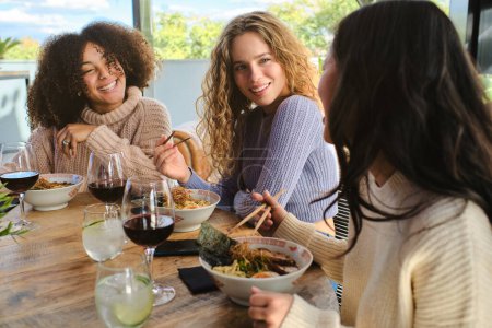 Foto de Jóvenes mujeres multirraciales en ropa casual sentados en la mesa con cuencos de ramen mientras pasan tiempo juntos - Imagen libre de derechos