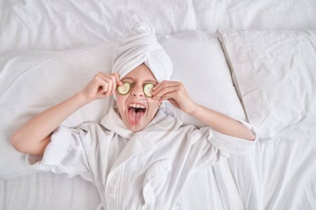 Téléchargez les photos : Vue du dessus de l'enfant excité en peignoir blanc et turban serviette montrant la langue et couvrant les yeux avec des tranches de concombre pendant les procédures de spa dans la chambre à coucher à la maison - en image libre de droit