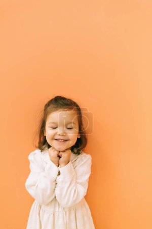 Téléchargez les photos : Mignon heureux tout-petit fille en robe blanche debout avec les yeux fermés près du mur orange et toucher le menton - en image libre de droit