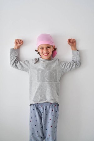 Photo for Cheerful cute boy in pajama and pink bandana smiling happily and looking at camera while standing against white wall and showing biceps - Royalty Free Image