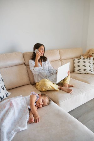 Foto de Cuerpo completo de madre joven que tiene conversación telefónica y computadora portátil de navegación mientras está sentado en el sofá cerca de la hija siesta y trabajando remotamente en la sala de estar en casa - Imagen libre de derechos