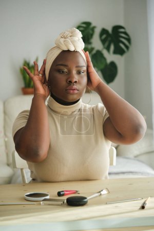 Téléchargez les photos : Headshot of serious young African American woman fixing beige turban and looking in mirror while sitting at dressing table in light bedroom - en image libre de droit