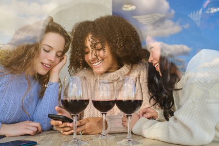 Foto de Diversas amigas positivas en suéteres con copas de vino tinto mirando la pantalla del teléfono inteligente - Imagen libre de derechos