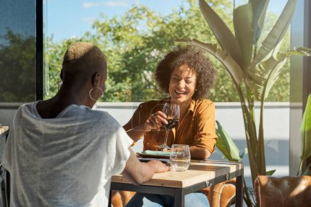 Téléchargez les photos : Rire amis féminines multiraciales en tenue décontractée assis à la table en bois et boire du vin rouge sur la terrasse du restaurant par une journée ensoleillée - en image libre de droit