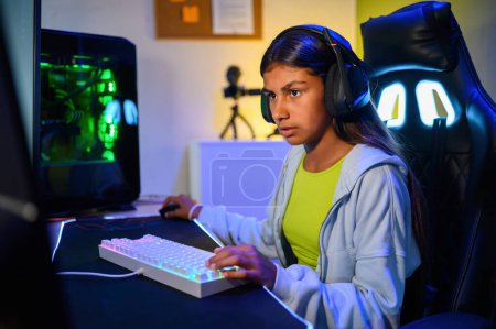 Téléchargez les photos : Vue latérale d'une jeune adolescente ethnique concentrée dans un casque regardant l'écran de l'ordinateur assis au bureau et jouant à des jeux vidéo près d'un PC de jeu - en image libre de droit