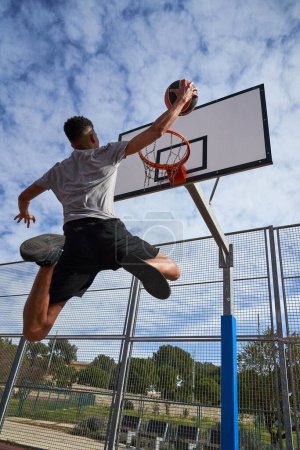 Foto de Vista trasera de ángulo bajo del jugador hispano de baloncesto saltando por encima del suelo y lanzando pelota mientras juega baloncesto en el campo de deportes - Imagen libre de derechos