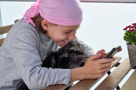 Photo for Side view of kid in pink headband sitting at wooden table with small dog and browsing mobile phone with smile - Royalty Free Image