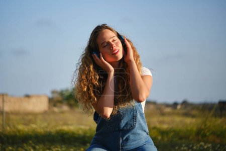Téléchargez les photos : Contenu jeune femme en combinaison profitant de la musique préférée dans les écouteurs sans fil tout en étant assis avec les yeux fermés sur prairie herbeuse dans la campagne estivale - en image libre de droit