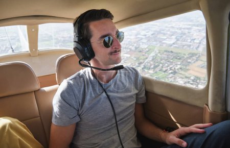 Foto de Joven viajero masculino positivo con bigote en traje casual y auriculares admirando el paisaje mientras está sentado en un pequeño avión moderno durante el vuelo en un día soleado - Imagen libre de derechos