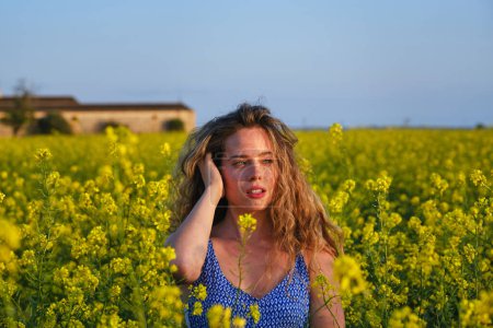 Téléchargez les photos : Superbe jeune femelle aux yeux bleus touchant les cheveux ondulés et regardant loin tout en se tenant debout sur une prairie fleurie avec des fleurs jaunes dans une campagne paisible - en image libre de droit