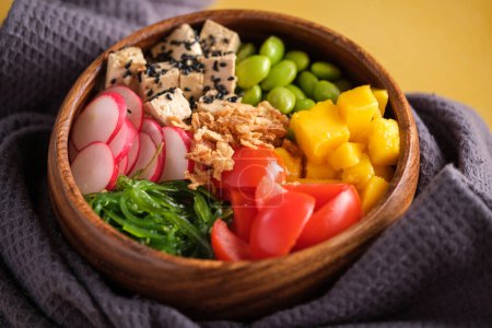 Photo for High angle of wooden bowl with fresh vegetables including sliced tomatoes radish tofu cheese and mango served with seaweed salad and edamame beans on towel on yellow table - Royalty Free Image