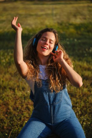Foto de Feliz joven hembra en traje de mezclilla bailando con el brazo levantado y cantando mientras escucha buena música en los auriculares y sentado con los ojos cerrados en el prado herboso en el campo - Imagen libre de derechos