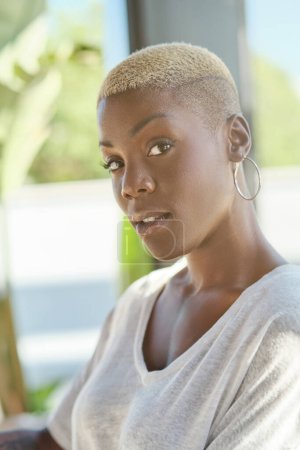 Téléchargez les photos : Portrait d'une jeune femme afro-américaine élégante et réfléchie aux cheveux teints courts et à la coiffure tendance portant un t-shirt blanc et des boucles d'oreilles regardant la caméra - en image libre de droit