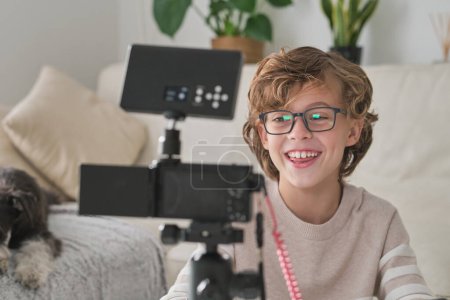 Téléchargez les photos : Garçon souriant en vêtements décontractés regardant caméra professionnelle et enregistrement vidéo pour blog dans le salon - en image libre de droit