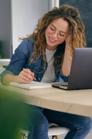 Téléchargez les photos : Jeune femme en vêtements décontractés assis à table avec ordinateur portable et travaillant sur le projet tout en prenant des notes dans le planificateur - en image libre de droit