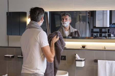 Foto de Vista posterior del hombre adulto en camiseta blanca mirando a la reflexión y haciendo la rutina de la mañana en el baño moderno en casa - Imagen libre de derechos
