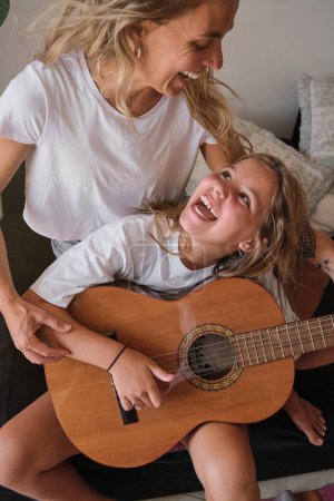 Foto de Foto vertical de una mujer rubia enseñando a una chica a tocar la guitarra mientras se miran riendo sentados en un sofá en una casa - Imagen libre de derechos